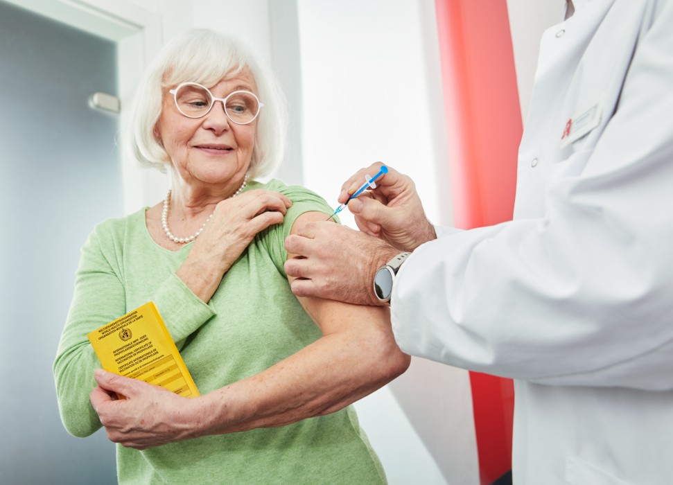 Ein Apotheker verabreicht einer älteren Dame, die ihren gelben Impfpass in der Hand hält, eine Impfung in den Oberarm