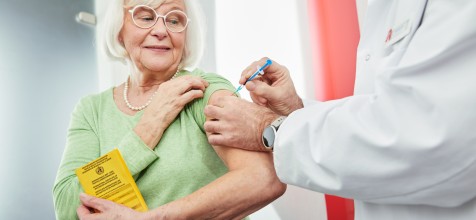 Ein Apotheker verabreicht einer älteren Dame, die ihren gelben Impfpass in der Hand hält, eine Impfung in den Oberarm